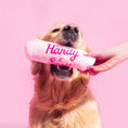 Charger l'image dans la visionneuse de la galerie, Patomia Bandit x Merci Handy pink plush lip gloss dog toy held in a golden retriever's mouth against a pink background
