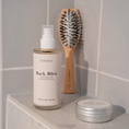 Charger l'image dans la visionneuse de la galerie, Patomia Bark Bliss leave-in dog conditioning spray bottle with a wooden brush and tin on a tiled bathroom shelf.

