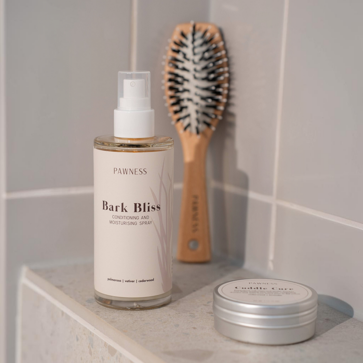 Patomia Bark Bliss leave-in dog conditioning spray bottle with a wooden brush and tin on a tiled bathroom shelf.