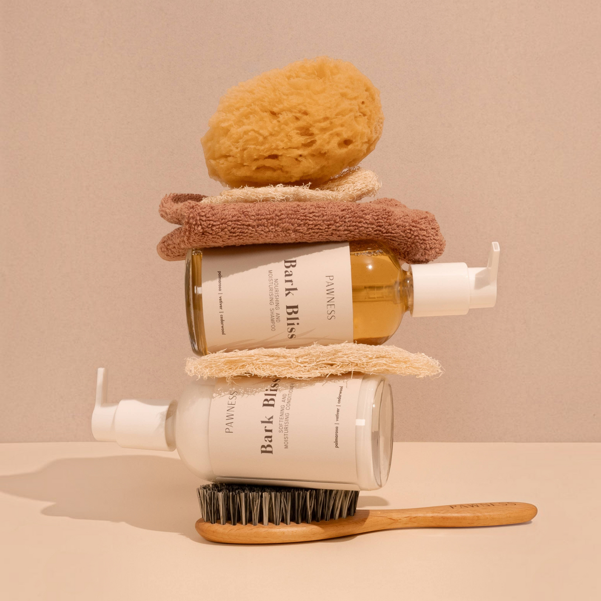 Patomia Bark Bliss dog shampoo and conditioner bottles stacked with a natural sponge, towel, and wooden grooming brush.