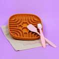 Charger l'image dans la visionneuse de la galerie, Patomia burnt orange silicone labyrinth lick mat with a pink spatula and toothbrush on a beige cloth against purple
