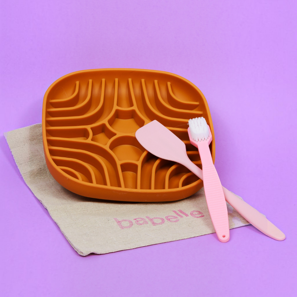 Patomia burnt orange silicone labyrinth lick mat with a pink spatula and toothbrush on a beige cloth against purple